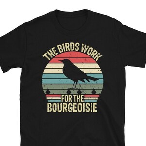 Puede incluir: Camiseta negra con un gráfico de estilo vintage de un pájaro frente a una puesta de sol. El texto dice "The Birds Work For The Bourgeoisie."
