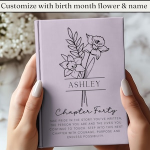 May include: A lavender journal with a floral design and the name "ASHLEY" printed on the cover. The text "Chapter Forty" and inspirational text are also visible. The journal is held by a person.