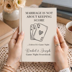 May include: A beige hardcover book titled Marriage Is Not About Keeping Score, featuring a line art illustration of three playing cards, the text ....Unless It's Game Night, and the personalized names Rachel & Joseph above Game Night Scorebook.