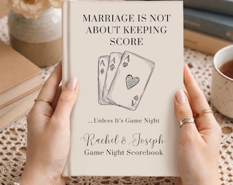Könnte beinhalten: Ein beigefarbenes Hardcover-Buch mit dem Titel Marriage Is Not About Keeping Score, einer Strichzeichnung von drei Spielkarten, dem Text ....Unless It's Game Night und den Namen Rachel & Joseph über Game Night Scorebook.