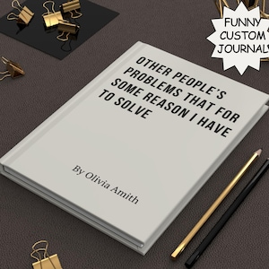 May include: A white journal with the text "OTHER PEOPLE'S PROBLEMS THAT FOR SOME REASON I HAVE TO SOLVE" and "By Olivia Amith". The journal is next to a gold and black pencil, and gold binder clips.
