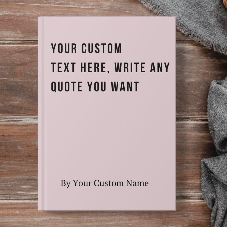 May include: A light pink journal with the text "YOUR CUSTOM TEXT HERE, WRITE ANY QUOTE YOU WANT" in black. The journal also includes the text "By Your Custom Name". The journal is closed and placed on a wooden surface.