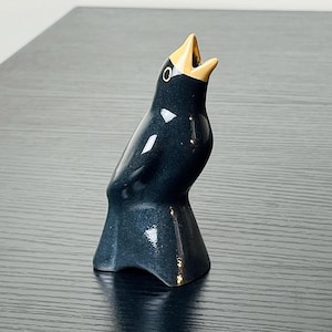May include: A ceramic figurine of a bird with a glossy black body and a golden beak. The bird is depicted with its beak open, as if singing. The figurine is placed on a dark wooden surface.