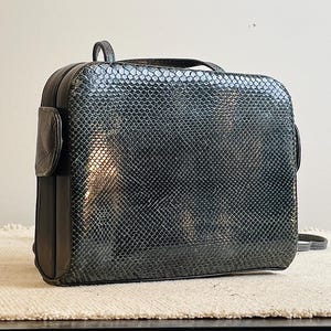 May include: A rectangular, dark gray handbag with a textured, snakeskin-like pattern. The bag has a small handle on top and a side pocket. The bag is sitting on a cream-colored surface.