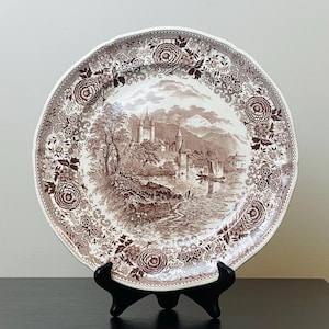 May include: A brown and white decorative plate featuring a scenic landscape with a castle, trees, and water. The plate has a detailed floral border and is displayed on a black stand.