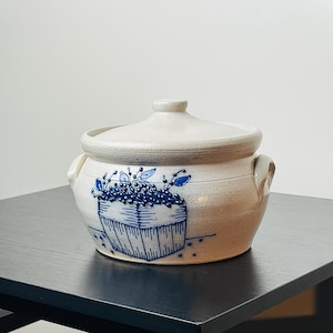 May include: A cream-colored ceramic pot with a lid, featuring a blue floral basket design. The pot has two small handles and is sitting on a black table. The pot is likely used for cooking or storage.