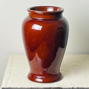 May include: A glossy, brown ceramic vase with a wide base and a slightly flared rim. The vase has a smooth, reflective surface and a classic, timeless design. It is a decorative item suitable for displaying flowers or as a standalone accent piece.
