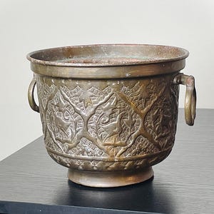 May include: Antique bronze planter with intricate embossed patterns. The planter features a wide, open top, two handles, and a decorative base. The embossed design includes stylized trees and animal figures. The planter is displayed on a black surface.