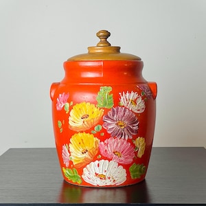May include: A vibrant orange ceramic jar with a wooden lid, adorned with hand-painted floral designs in yellow, pink, purple, and white. The jar has a glossy finish and is set on a dark surface.
