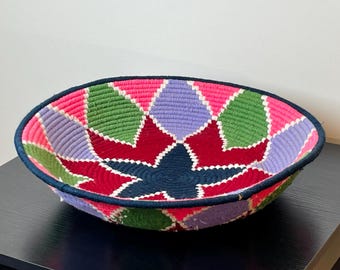 May include: A colorful, woven basket with a starburst design. The oval-shaped basket features a navy blue rim and a pattern of red, pink, green, and purple geometric shapes. The basket is placed on a black table.