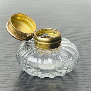May include: A clear glass inkwell with a ribbed design and a hinged brass lid. The lid is open, revealing the interior. The inkwell has a round shape and sits on a dark surface.