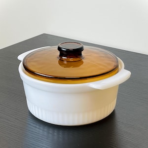 May include: A white casserole dish with a clear amber-colored glass lid and a black knob. The dish has two white handles and a ribbed design around the base. A kitchenware item.