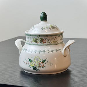 May include: A white ceramic sugar bowl with a lid, featuring floral patterns in yellow, orange, and green. The bowl has two handles and a green knob on the lid. The design includes a floral basket motif.