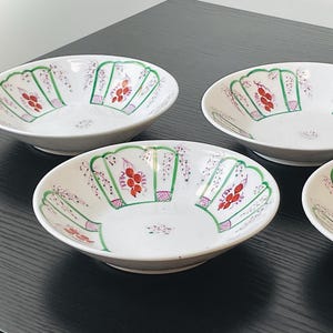 May include: Four white ceramic bowls with a scalloped rim design. Each bowl features a hand-painted pattern of red floral clusters, green leaves, and pink accents. The bowls are arranged on a dark surface.