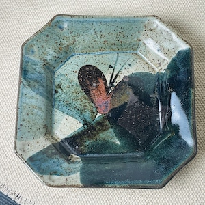 May include: Square ceramic plate with an octagonal shape. The plate features a speckled teal and blue glaze with a dark brown and orange abstract design in the center. The plate is a decorative piece.