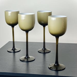 May include: Four olive-green wine glasses with a rounded bowl and a long, slender stem. The glasses have a dark base and stem, and a white interior. The glasses are arranged on a dark surface.