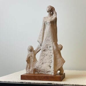 May include: A beige sculpture of a woman and child. The woman is wearing a long dress and the child is holding her hand. The sculpture is mounted on a rectangular wooden base. The figures have a textured surface.