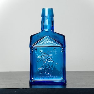 May include: A vibrant blue glass bottle with a rectangular body and a detailed embossed design. The bottle features a rider on a horse and architectural elements. The bottle is approximately 8 inches tall.