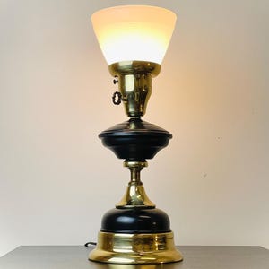 May include: An antique table lamp with a brass base and accents, featuring a black central body and a frosted glass shade. The lamp is illuminated, casting a warm glow. The design is reminiscent of vintage lighting.