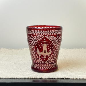 May include: A ruby red glass with a white lighthouse design, surrounded by a decorative floral pattern. The glass has a slightly flared shape and is likely used for drinking. The design is etched into the glass.
