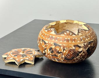 May include: A decorative, round bowl with a star-shaped lid. The bowl and lid are brown with intricate, carved designs in shades of brown and black. The bowl sits on a black surface.