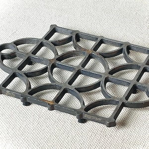 May include: A dark gray, cast iron trivet with a geometric design. The trivet features a pattern of interlocking circles, triangles, and squares. It has a rectangular shape with rounded corners and small feet.