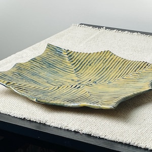 May include: A handmade ceramic leaf-shaped dish with a textured surface. The dish features a yellow and blue glaze, with a ribbed pattern mimicking leaf veins. The edges are slightly raised, and the dish sits on a neutral-colored linen cloth.