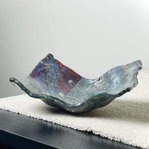 May include: A handcrafted ceramic bowl with a unique, organic shape. The bowl features a blend of blue, red, and gray hues, with a textured surface and a wavy rim. The bowl is approximately 12 inches long and 8 inches wide.