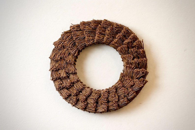 May include: A circular wreath made of brown twigs, arranged in layers to create a textured, layered effect. The wreath is a decorative item, suitable for seasonal or year-round display. The wreath is set against a plain white background.