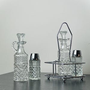 May include: A clear glass cruet set with a diamond pattern, including two cruets with handles and stoppers, and two shakers with silver-colored tops. One set is in a chrome carrier.