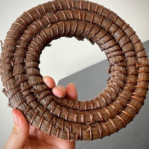 May include: A circular, woven wreath made of brown natural fibers and copper wire. The wreath is held in a hand and has a hollow center. The woven design is tightly packed and creates a textured surface.