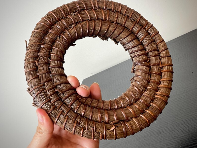 May include: A circular, woven wreath made of brown natural fibers and copper wire. The wreath is held in a hand and has a hollow center. The woven design is tightly packed and creates a textured surface.