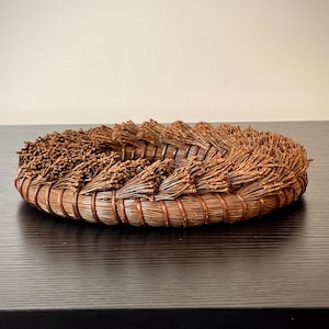 May include: A handcrafted, circular basket made of natural brown materials. The basket features a woven base with bundles of dried plant material arranged around the rim. The design is rustic and organic, suitable for home decor or storage.