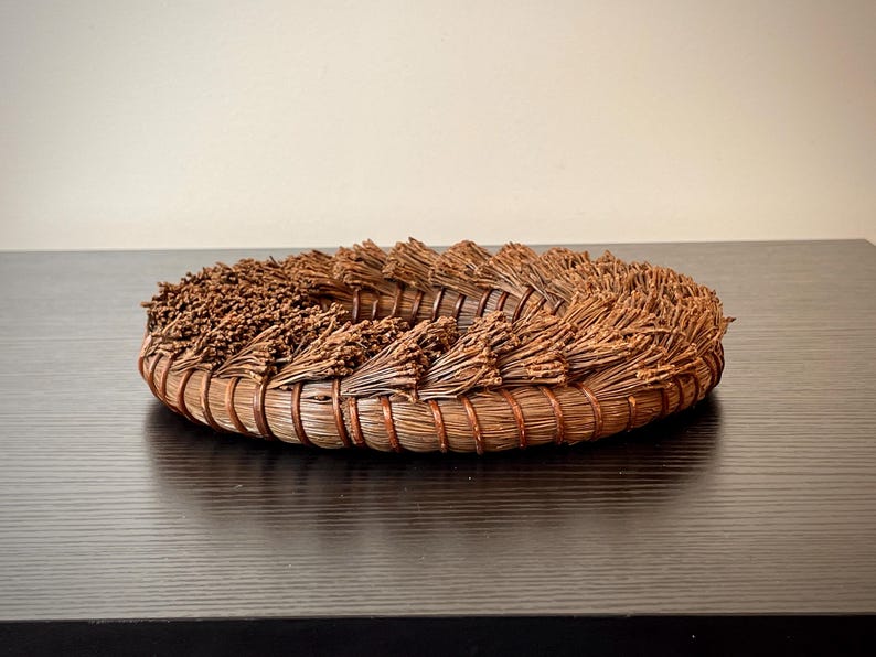 May include: A handcrafted, circular basket made of natural brown materials. The basket features a woven base with bundles of dried plant material arranged around the rim. The design is rustic and organic, suitable for home decor or storage.