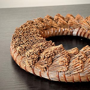 May include: A circular, handcrafted wreath made of natural brown materials. The wreath features a woven base with bundles of dried, textured elements arranged around the circumference. The design suggests a rustic, organic aesthetic, suitable for home decor.