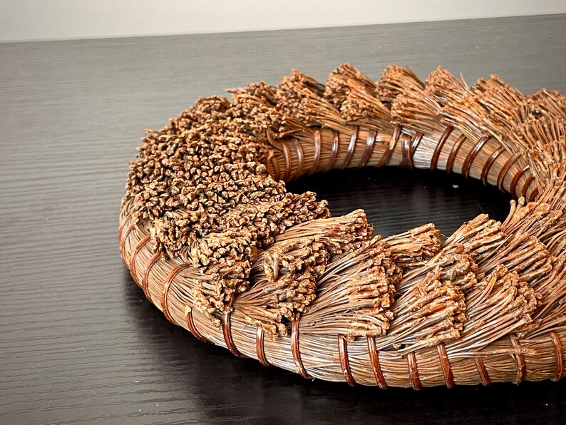 May include: A circular, handcrafted wreath made of natural brown materials. The wreath features a woven base with bundles of dried, textured elements arranged around the circumference. The design suggests a rustic, organic aesthetic, suitable for home decor.