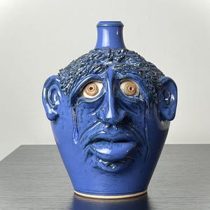 May include: A cobalt blue ceramic jug with a sculpted face. The face has large, expressive eyes, a downturned mouth, and a tear rolling down the cheek. The jug has a textured, hair-like design around the top.