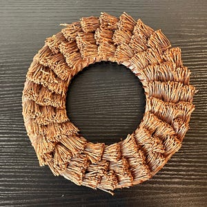 May include: A circular wreath made of layered, dried, brown plant material. The wreath has a central opening and is arranged in a radial pattern. The natural-toned wreath is suitable for home decor.