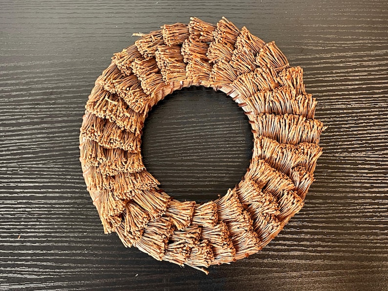 May include: A circular wreath made of layered, dried, brown plant material. The wreath has a central opening and is arranged in a radial pattern. The natural-toned wreath is suitable for home decor.