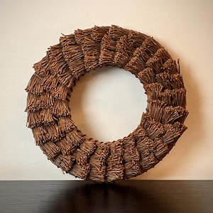 May include: A circular wreath made of brown twigs. The wreath is a decorative item, likely for seasonal or home decor. The twigs are arranged in a layered pattern, creating a textured, natural appearance. The wreath is displayed against a neutral background.