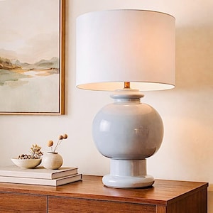 May include: A light blue ceramic table lamp with a white lampshade. The lamp has a round base and body, and a cylindrical shade. The lamp is on a wooden table. A painting and small decorative items are in the background.