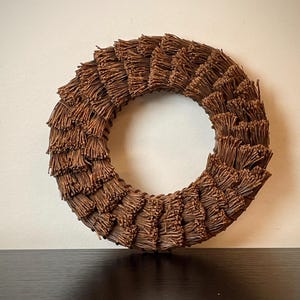 May include: A circular brown wreath made of bundled, textured twigs. The wreath is displayed against a neutral background, showcasing its natural, rustic aesthetic. The wreath's design is symmetrical, with the twigs radiating outwards.