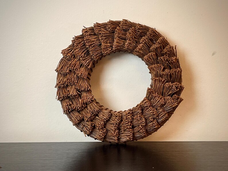 May include: A circular brown wreath made of bundled, textured twigs. The wreath is displayed against a neutral background, showcasing its natural, rustic aesthetic. The wreath's design is symmetrical, with the twigs radiating outwards.