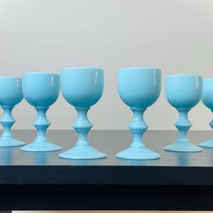 May include: Six light blue goblets with a rounded bowl and a detailed stem. The goblets are arranged in a row on a dark surface, with a plain white background. The glassware is likely for serving beverages.