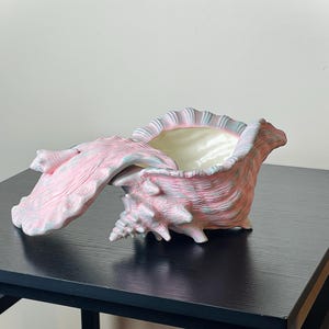 May include: A seashell-shaped decorative box with a lid. The box is painted in shades of pink, blue, and white, with a creamy interior. The lid is partially open, revealing the inside. The box is sitting on a black table.