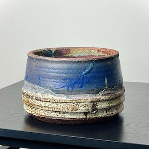 May include: A handmade ceramic bowl with a wide, open top. The bowl features a layered design with shades of blue, brown, and cream. The exterior has a textured, rough appearance, and the interior is a darker brown.