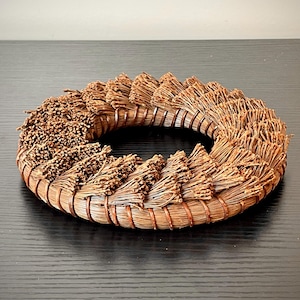 May include: A circular, woven wreath made of dried, brown plant material. The wreath is arranged in a spiral pattern, with the plant material radiating outwards from the center. The wreath is sitting on a dark surface.