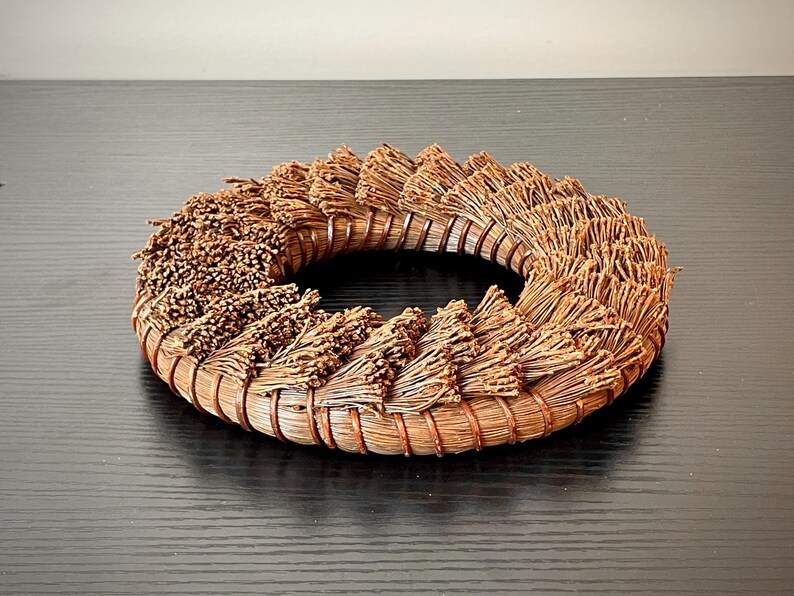 May include: A circular, woven wreath made of dried, brown plant material. The wreath is arranged in a spiral pattern, with the plant material radiating outwards from the center. The wreath is sitting on a dark surface.
