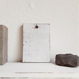May include: White wooden sign with a distressed finish and a small hole in the center. The sign is sitting on a white surface.
