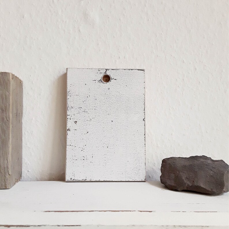 May include: White wooden sign with a distressed finish and a small hole in the center. The sign is sitting on a white surface.
